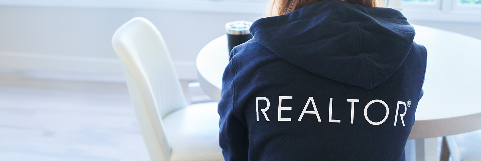 Image of Woman sitting at a kitchen table with her back to the camera, wearing a navy sweatshirt that has the "REALTOR®" printed on it. 